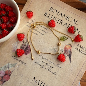 Puede incluir: Un brazalete de cadena dorada con un dije de frambuesa roja y un dije de hoja verde. El brazalete está sobre un trozo de papel con el texto "Botanical Illustration" y "Parus Major" impreso en él.