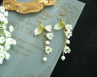 May include: A pair of gold dangle earrings with white flower charms and pearl accents. The flowers are green and white and resemble lily of the valley.