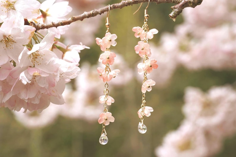 May include: Delicate dangle earrings featuring cascading pink flower charms and sparkling crystal teardrops. The earrings are suspended from gold-colored hooks and chains, creating a graceful, feminine accessory. The earrings are shown against a blurred background of pink blossoms.