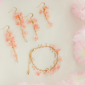 May include: A jewelry set featuring delicate pink flower earrings and a bracelet with gold-tone accents. The earrings dangle with clear crystal accents. The bracelet and earrings are arranged on a white surface with pink floral accents.