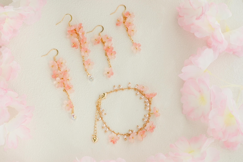 May include: A jewelry set featuring delicate pink flower earrings and a bracelet with gold-tone accents. The earrings dangle with clear crystal accents. The bracelet and earrings are arranged on a white surface with pink floral accents.
