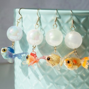 May include: Four pairs of dangle earrings with iridescent pearl-like beads and gold fish charms. The fish charms are in blue, orange, gold, and gold with orange accents.