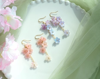 Könnte beinhalten: Drei Paar Hängeohrringe mit zarten Blumenanhängern. Die Ohrringe sind aus pastellrosa, hellblau und lila Blumen gefertigt.