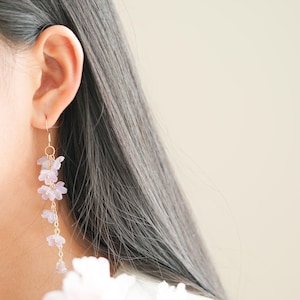 May include: A close-up of a person's ear wearing a pair of long, dangling earrings. The earrings feature delicate, light purple flower clusters suspended from gold-colored hooks and chains. The background is a soft, neutral color, highlighting the jewelry.