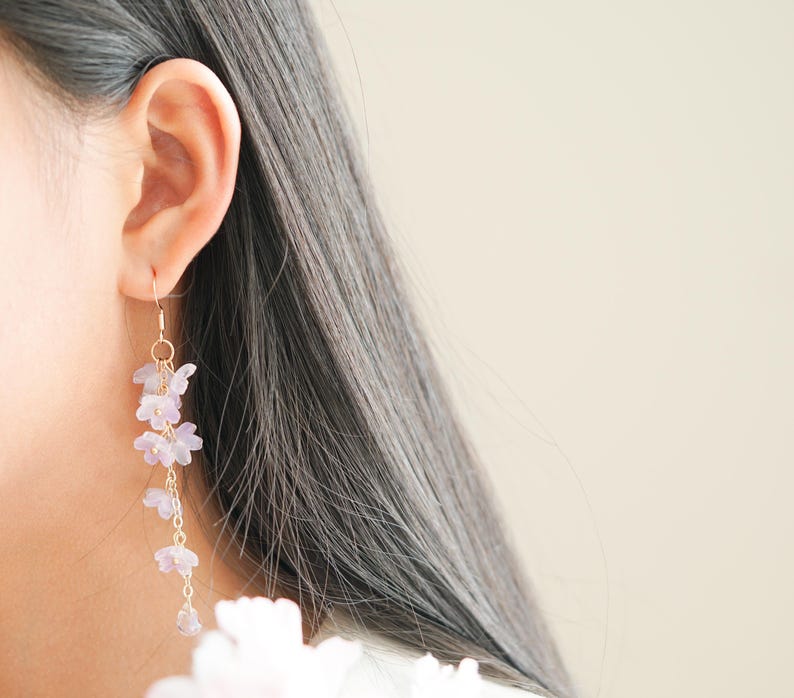 May include: A close-up of a person's ear wearing a pair of long, dangling earrings. The earrings feature delicate, light purple flower clusters suspended from gold-colored hooks and chains. The background is a soft, neutral color, highlighting the jewelry.