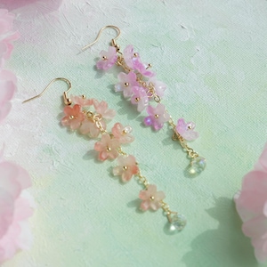 May include: A pair of delicate floral earrings. Each earring features a cascade of pink and peach-colored flower charms suspended from gold-colored hooks. Clear crystal-like beads dangle at the bottom of each earring, adding a touch of sparkle.
