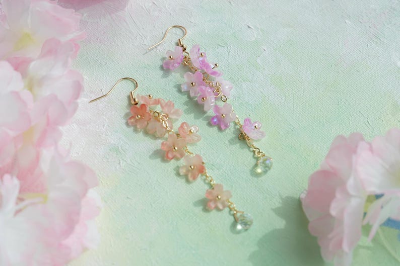 May include: A pair of delicate floral earrings. Each earring features a cascade of pink and peach-colored flower charms suspended from gold-colored hooks. Clear crystal-like beads dangle at the bottom of each earring, adding a touch of sparkle.