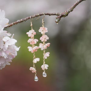 May include: Delicate dangle earrings featuring cascading pink flower charms and clear teardrop-shaped crystals. The earrings are suspended from gold-colored hooks and chains. The earrings are displayed on a tree branch, with a blurred floral background.