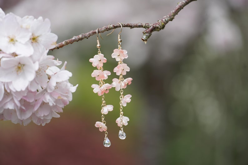 May include: Delicate dangle earrings featuring cascading pink flower charms and clear teardrop-shaped crystals. The earrings are suspended from gold-colored hooks and chains. The earrings are displayed on a tree branch, with a blurred floral background.