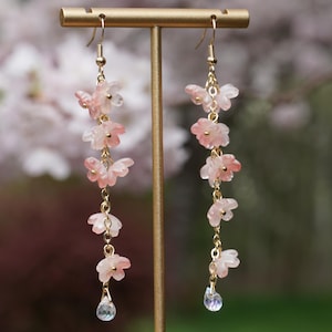 May include: Dangling earrings featuring delicate pink flower charms and a clear teardrop-shaped crystal. The earrings are suspended from gold-colored hooks and chains. The floral charms are arranged in a cascading design, creating an elegant and feminine accessory.