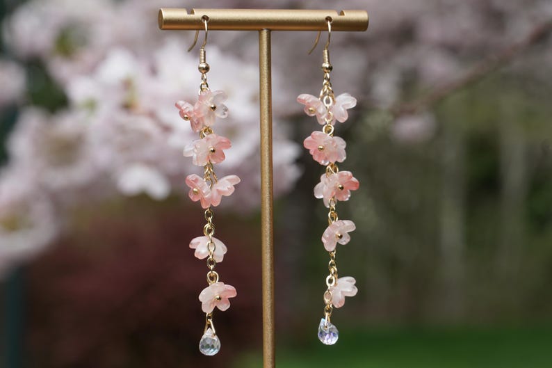 May include: Dangling earrings featuring delicate pink flower charms and a clear teardrop-shaped crystal. The earrings are suspended from gold-colored hooks and chains. The floral charms are arranged in a cascading design, creating an elegant and feminine accessory.