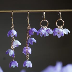 May include: A pair of dangle earrings with purple flower charms. The earrings are made with gold-toned metal and feature a delicate chain with multiple flower charms.