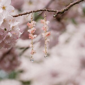 May include: Delicate gold-tone earrings featuring cascading pink flower charms and a small, clear crystal at the bottom. The earrings are suspended from a branch with soft pink blossoms in the background, creating a romantic and feminine aesthetic.