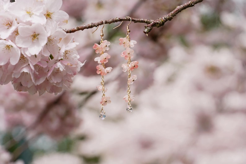 May include: Delicate gold-tone earrings featuring cascading pink flower charms and a small, clear crystal at the bottom. The earrings are suspended from a branch with soft pink blossoms in the background, creating a romantic and feminine aesthetic.