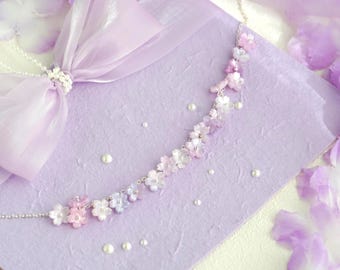 May include: A delicate floral necklace with pink, white, and lavender blossoms. The necklace is set on a lavender background with scattered pearls and a matching ribbon. The design evokes a sense of elegance and femininity.