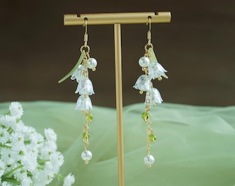 May include: A pair of dangle earrings with white lily of the valley flowers, green beads, and pearl accents. The earrings are hanging from a gold stand.