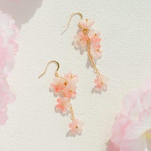 May include: A pair of delicate gold-tone earrings featuring clusters of pink and white flower charms. The earrings have a cascading design with small dangling flowers. The background is a soft white with pink floral accents.