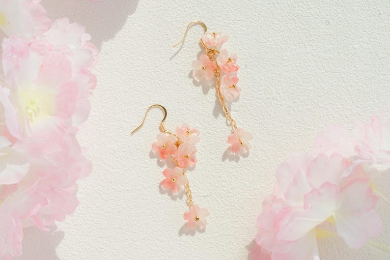 May include: A pair of delicate gold-tone earrings featuring clusters of pink and white flower charms. The earrings have a cascading design with small dangling flowers. The background is a soft white with pink floral accents.