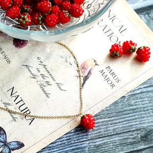 May include: A gold chain necklace with a red raspberry charm. The necklace is lying on a page from a book with text about birds and nature. There are also red raspberry charms on the page.