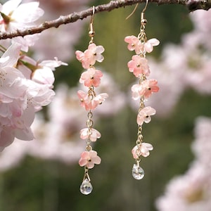 May include: Delicate dangle earrings featuring pink flower charms and clear crystal teardrops. The earrings are suspended from gold-colored chains and hooks. The background includes soft-focus pink blossoms and a tree branch.