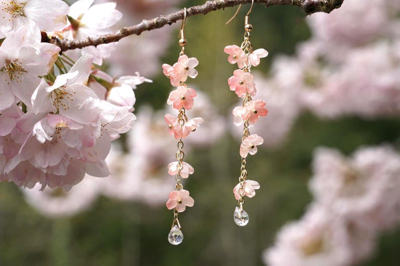 May include: Delicate dangle earrings featuring pink flower charms and clear crystal teardrops. The earrings are suspended from gold-colored chains and hooks. The background includes soft-focus pink blossoms and a tree branch.