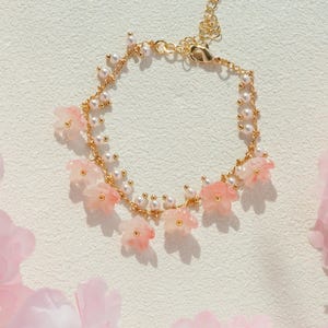 May include: A delicate gold-toned bracelet adorned with small pink flower charms and white pearls. The bracelet is set against a soft, textured background, with pink flowers in the foreground.