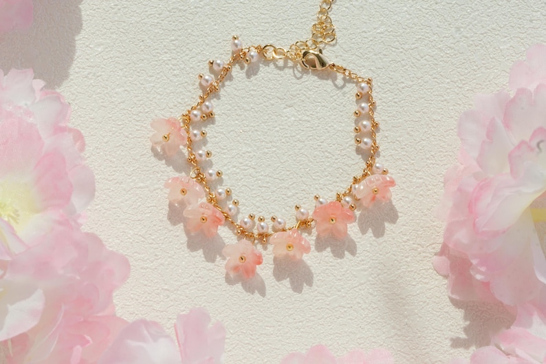May include: A delicate gold-toned bracelet adorned with small pink flower charms and white pearls. The bracelet is set against a soft, textured background, with pink flowers in the foreground.