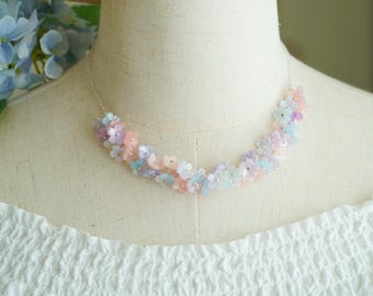 May include: A delicate floral necklace composed of small, pastel-colored flowers in shades of pink, blue, and lavender. The necklace is strung on a thin silver chain and rests on a mannequin. The flowers are clustered together to create a garland effect.