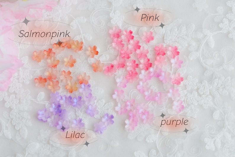 May include: A collection of small, delicate flower embellishments in shades of salmon, pink, and lilac. The image includes text labels: "Salmonpink", "Pink", "Lilac", and "purple". The flowers are scattered on a white, lacy background.