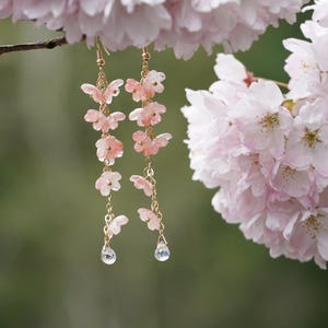 May include: Delicate gold-tone earrings with cascading pink butterfly charms and teardrop-shaped crystal pendants. The earrings are suspended from a branch with soft pink cherry blossoms in the background, creating a romantic and feminine aesthetic.