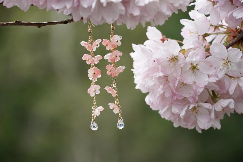 May include: Delicate gold-tone earrings with cascading pink butterfly charms and teardrop-shaped crystal pendants. The earrings are suspended from a branch with soft pink cherry blossoms in the background, creating a romantic and feminine aesthetic.