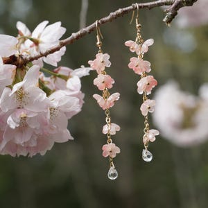 May include: Delicate gold earrings with pink flower and butterfly charms, accented with clear crystal teardrops. The earrings hang from a branch with soft pink cherry blossoms, creating a nature-inspired aesthetic.