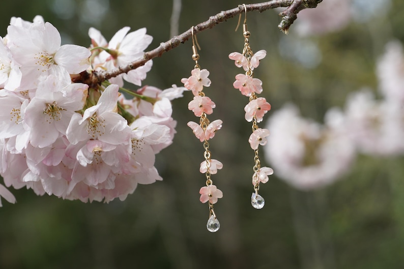 May include: Delicate gold earrings with pink flower and butterfly charms, accented with clear crystal teardrops. The earrings hang from a branch with soft pink cherry blossoms, creating a nature-inspired aesthetic.