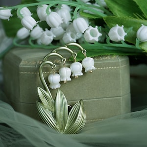 May include: A gold-toned brooch in the shape of a lily of the valley flower. The brooch has white enamel flowers and green enamel leaves.