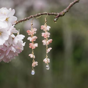 May include: Dangling earrings featuring delicate pink flower charms suspended from gold-colored hooks. Each earring has multiple flower charms and a small, clear crystal at the bottom. The earrings are displayed on a tree branch with a blurred background of pink flowers.
