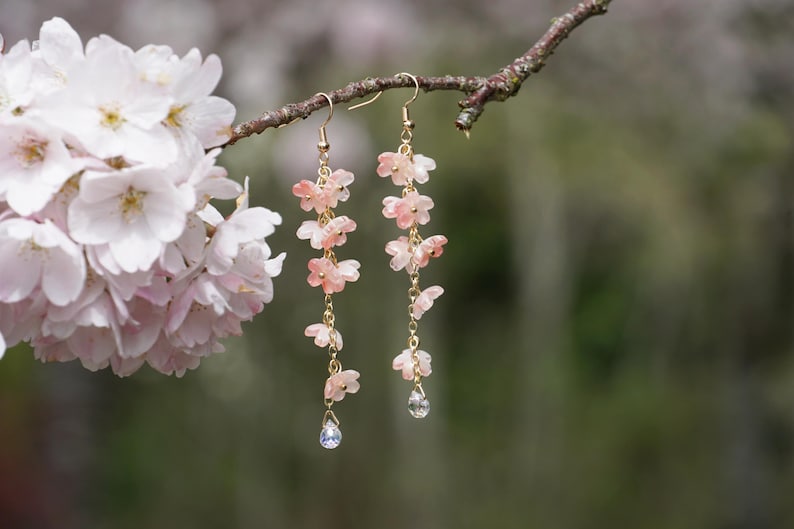 May include: Dangling earrings featuring delicate pink flower charms suspended from gold-colored hooks. Each earring has multiple flower charms and a small, clear crystal at the bottom. The earrings are displayed on a tree branch with a blurred background of pink flowers.