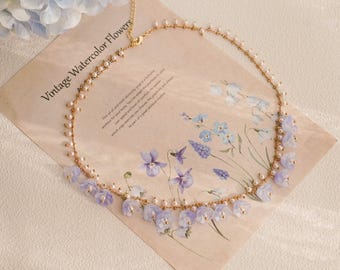 May include: A delicate gold-tone necklace featuring small, light purple flower pendants and tiny pearls. The necklace is displayed on a vintage-style paper with watercolor floral illustrations and the text "Vintage Watercolor Flowers".