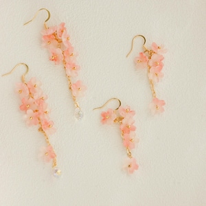 May include: Four pairs of pink floral earrings with gold-colored hooks and chains. The earrings feature delicate, cascading flower clusters and small, clear crystal accents. The earrings are set against a soft, off-white background.