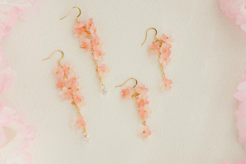 May include: Four pairs of pink floral earrings with gold-colored hooks and chains. The earrings feature delicate, cascading flower clusters and small, clear crystal accents. The earrings are set against a soft, off-white background.