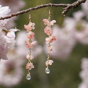 May include: Dangling earrings featuring delicate pink flower charms and a clear crystal teardrop. The earrings are suspended from gold-colored hooks and chains, creating a graceful, floral design. The background is a soft, blurred image of pink blossoms.