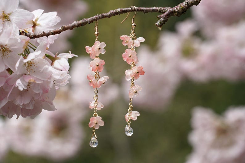 May include: Dangling earrings featuring delicate pink flower charms and a clear crystal teardrop. The earrings are suspended from gold-colored hooks and chains, creating a graceful, floral design. The background is a soft, blurred image of pink blossoms.