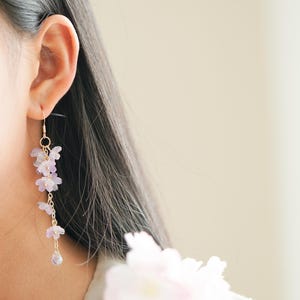 May include: A delicate gold-toned earring with a cascading floral design. The earring features small, light purple flowers and a teardrop-shaped charm at the bottom. The earring is worn on the ear of a person with dark hair.