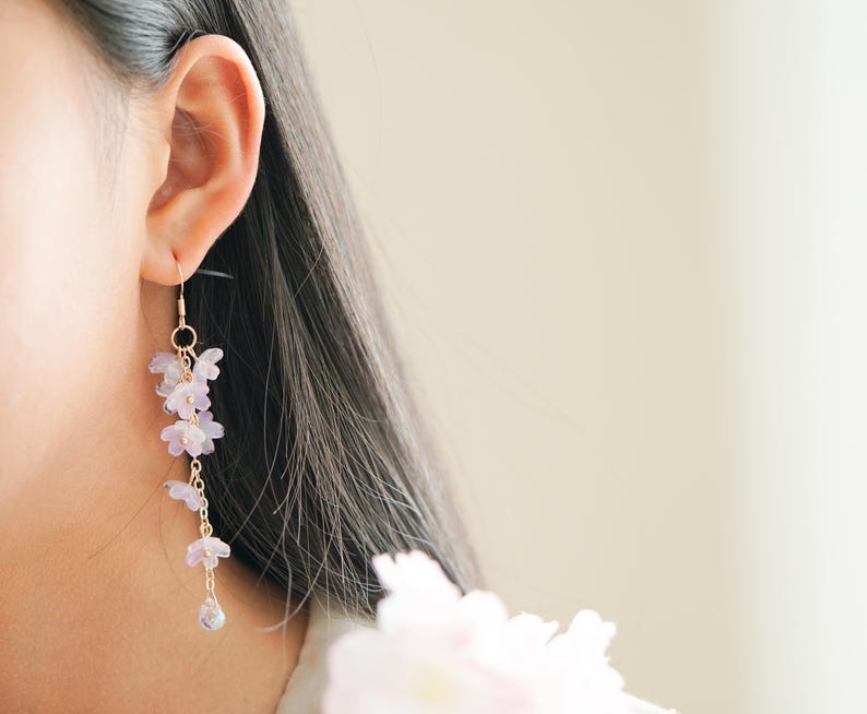 May include: A delicate gold-toned earring with a cascading floral design. The earring features small, light purple flowers and a teardrop-shaped charm at the bottom. The earring is worn on the ear of a person with dark hair.