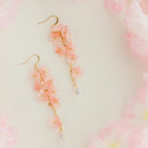 May include: A pair of delicate, gold-toned earrings featuring cascading clusters of small, light pink flowers. Each earring has a small, clear bead at the bottom. The earrings are set against a soft, textured white background, with pink floral accents.
