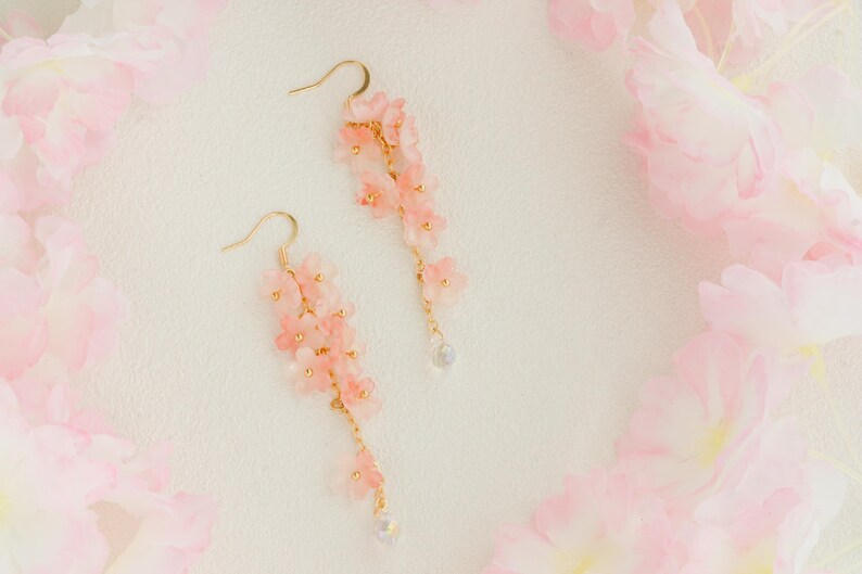 May include: A pair of delicate, gold-toned earrings featuring cascading clusters of small, light pink flowers. Each earring has a small, clear bead at the bottom. The earrings are set against a soft, textured white background, with pink floral accents.