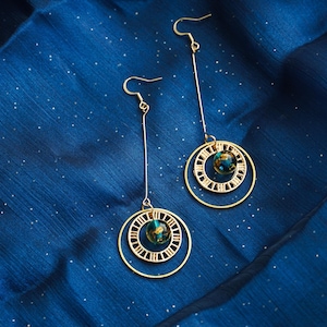 Peut inclure: Boucles d'oreilles pendantes dorées avec un thème céleste. Chaque boucle d'oreille est composée d'une longue barre fine reliée à un pendentif à double cercle avec des chiffres romains et un petit globe marbré. Fond bleu foncé texturé.