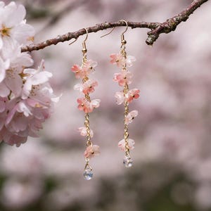 May include: Dangling earrings with delicate pink flower charms and clear crystal accents. The earrings are suspended from gold-colored hooks and chains, with a blurred background of pink blossoms.