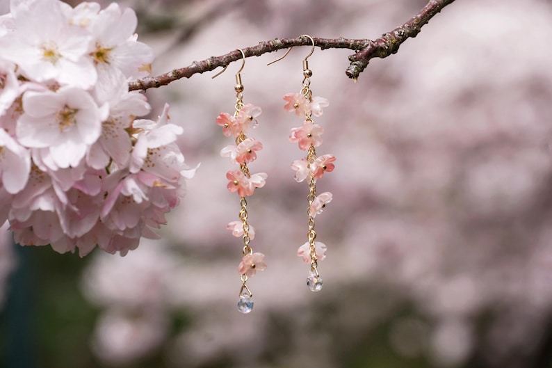 May include: Dangling earrings with delicate pink flower charms and clear crystal accents. The earrings are suspended from gold-colored hooks and chains, with a blurred background of pink blossoms.