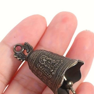 May include: A small, ornate, bronze-colored bell with a decorative design and a loop at the top. The bell is held in a hand.