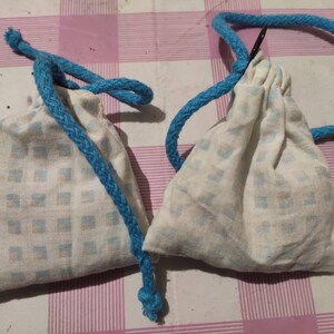 Può includere: Due piccole borse di stoffa bianca con coulisse, con motivi a quadri blu e bianchi e coulisse blu. Le borse sono su una superficie a quadri rosa e bianchi.